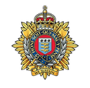 The cap badge of the Royal Logistic Corps: a crowned garter inscribed 'Honi Soit Qui Mal Y Pense' surrounding the corps shield, flanked by laurel and a gold sunburst, with the motto 'We Sustain' beneath.